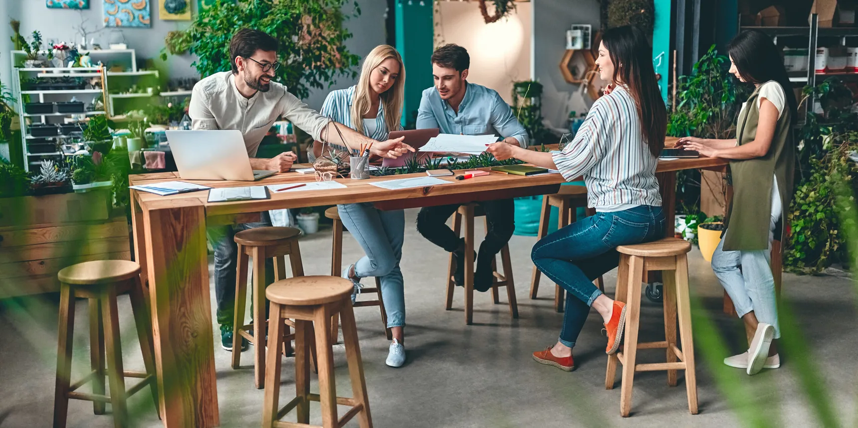 Cinq personnes travaillent autour d'une table avec ordinateur portable, documents et plantes