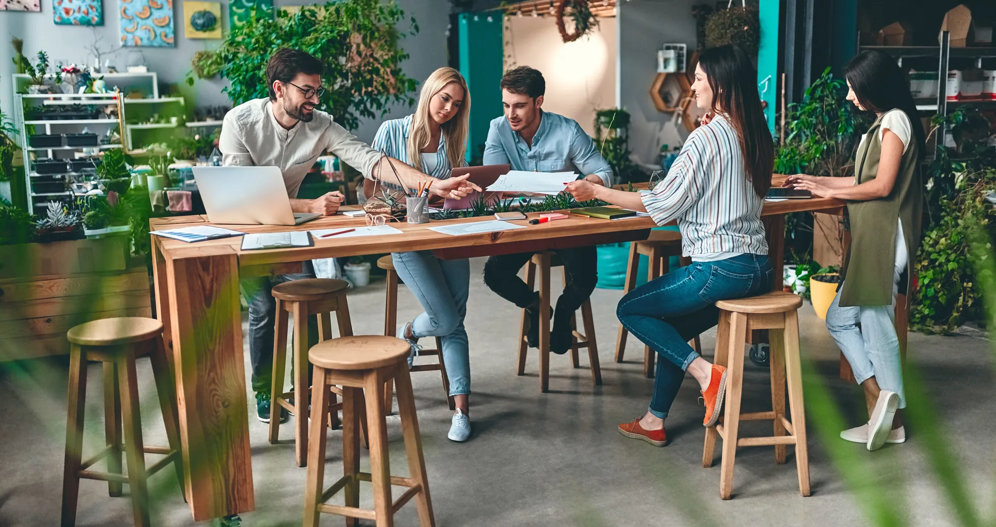 Cinq personnes travaillent autour d'une table avec ordinateur portable, documents et plantes
