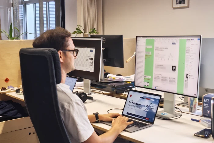 Designer de profil à son poste de travail, devant un grand écran de maquette et un ordinateur portable ouvert sur une page web