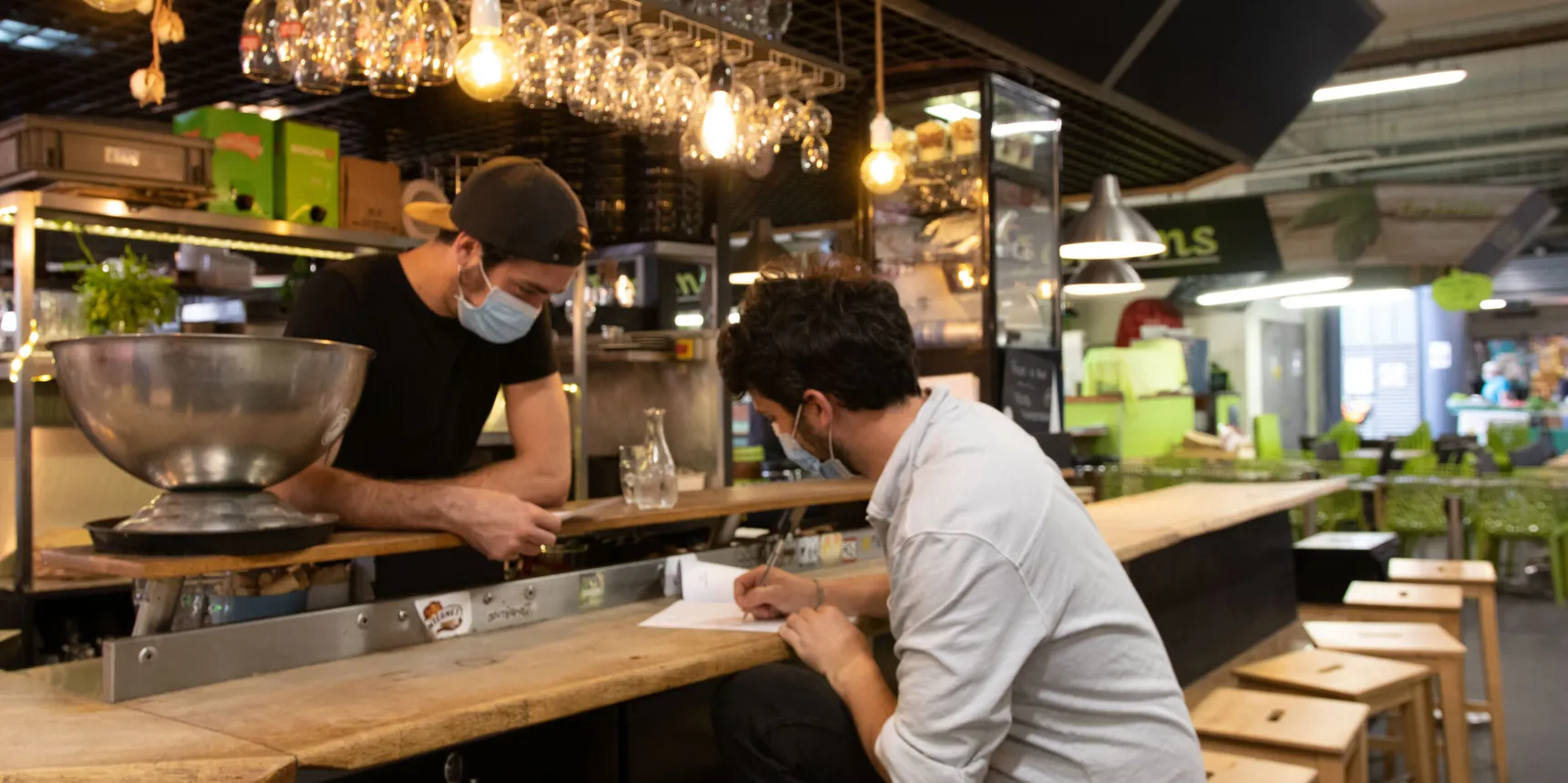 Two masked people speak across the counter of an establishment