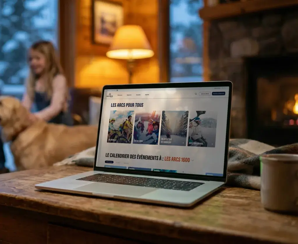 Laptop on a wooden table, showing the webpage of a mountain resort titled "Les Arcs for everyone". In the background, a cosy chalet living room with a stone fireplace and a large window opening onto a snowy landscape. Two children and a Golden Retriever are blurred in the background.