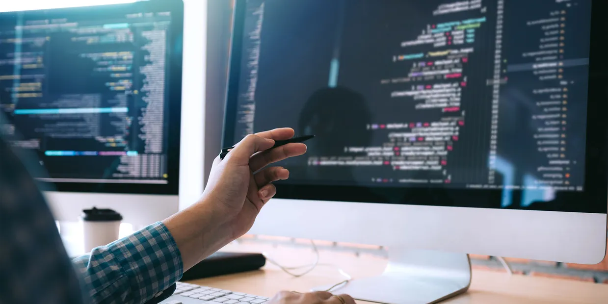 Hand pointing at code displayed across two computer monitors