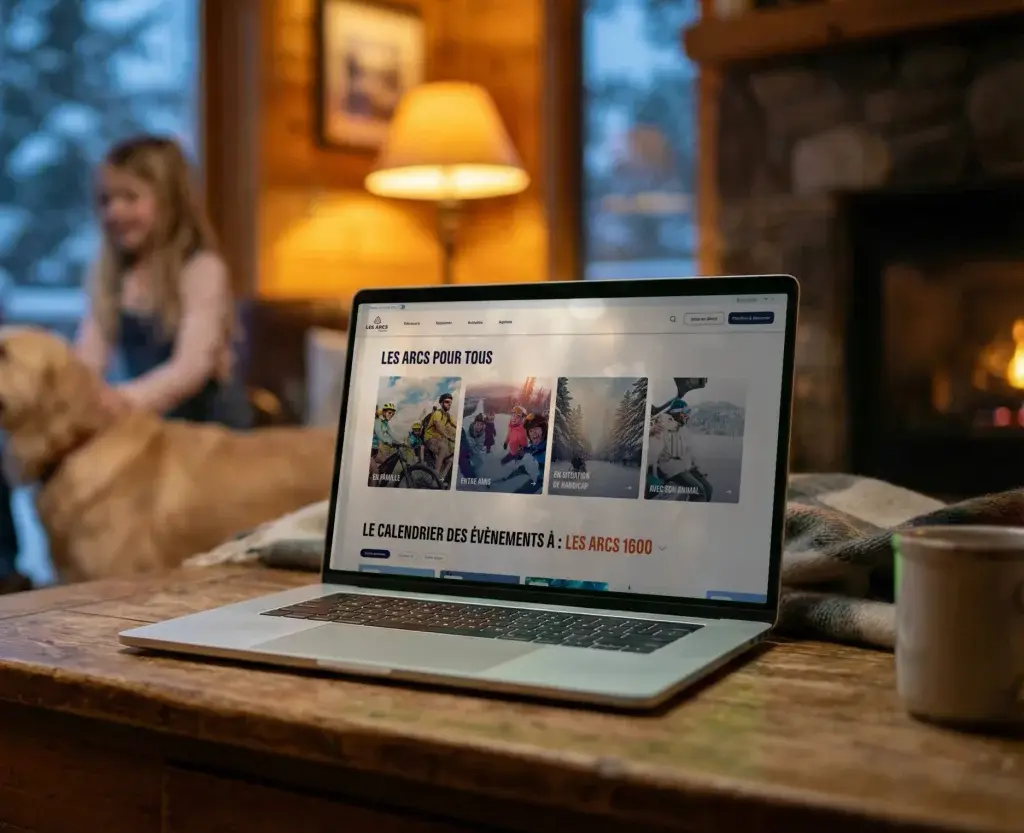 Laptop on a wooden table, showing the webpage of a mountain resort titled "Les Arcs for everyone". In the background, a cosy chalet living room with a stone fireplace and a large window opening onto a snowy landscape. Two children and a Golden Retriever are blurred in the background.
