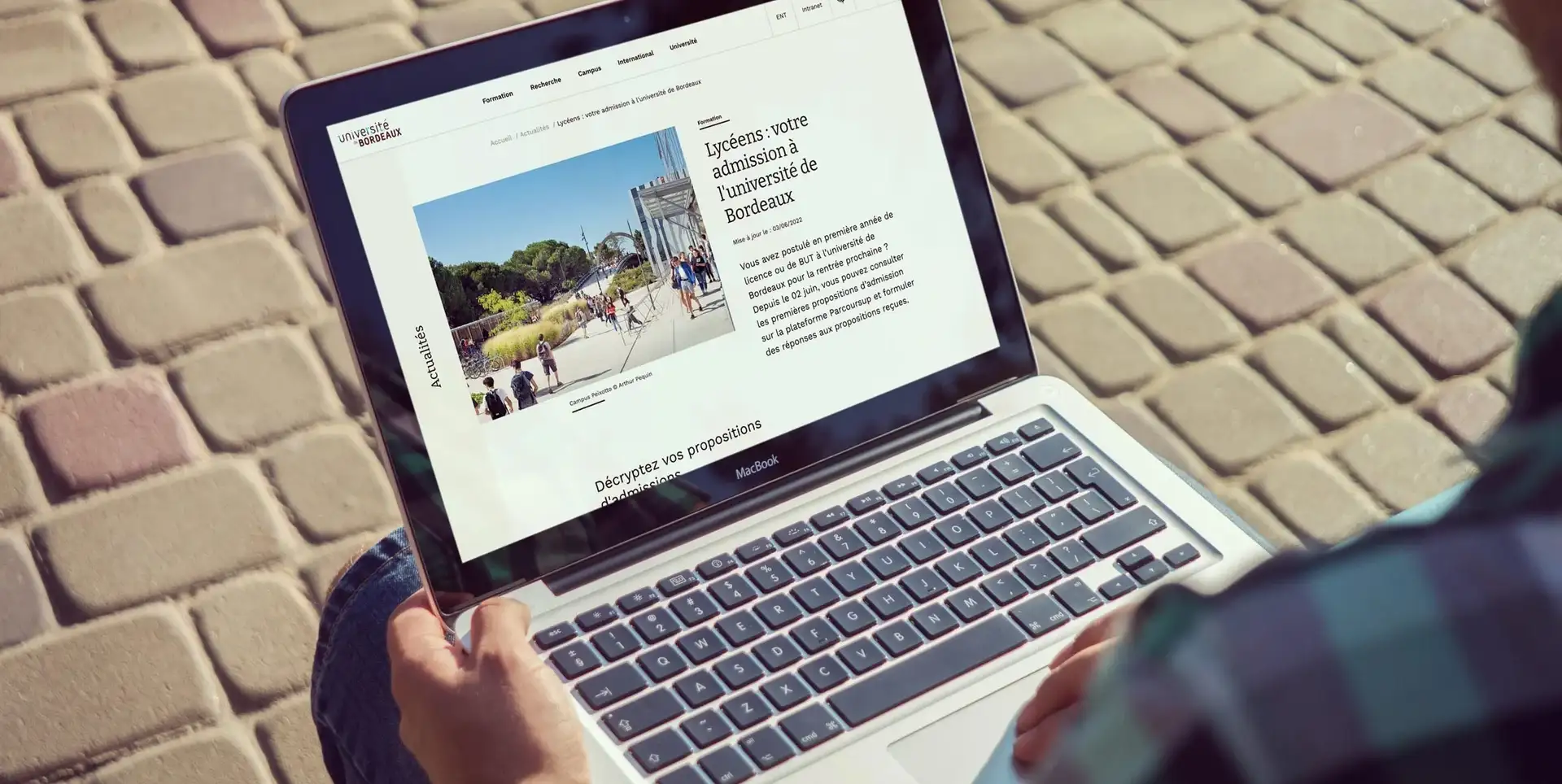 Personne assise sur des pavés avec un ordinateur affichant l’article « Lycéens : votre admission à l’université de Bordeaux »