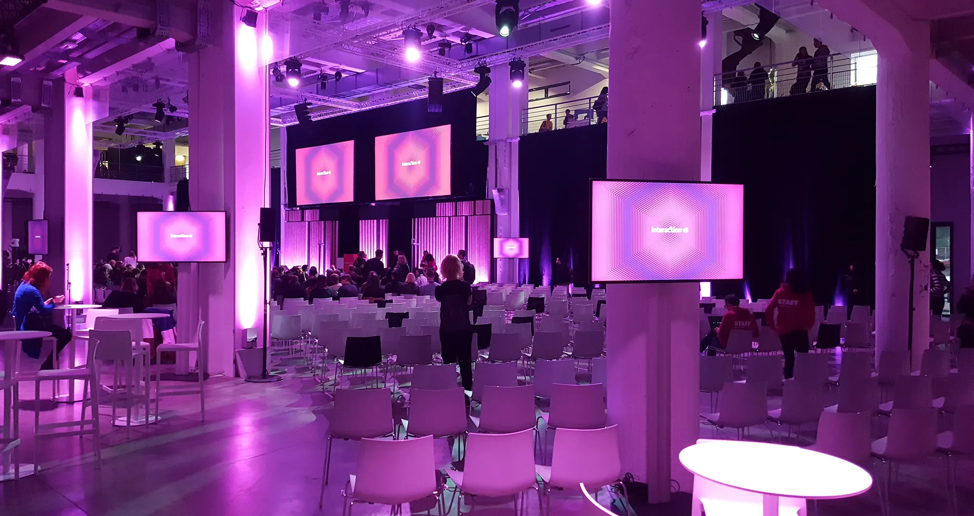 Conference hall lit in purple with rows of chairs and numerous screens