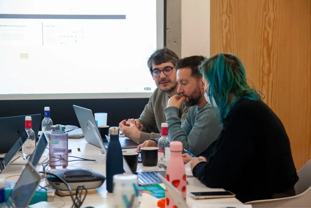 Three people are sitting around a meeting table, in front of a projection screen displaying a user-flow diagram. They are in mid-discussion, visibly focused on an RFP. The table is cluttered with water bottles, flasks, cups, a laptop and other work equipment