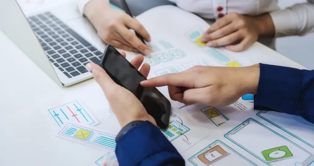 Trois personnes examinent un téléphone et des maquettes d'interfaces disposées sur une table