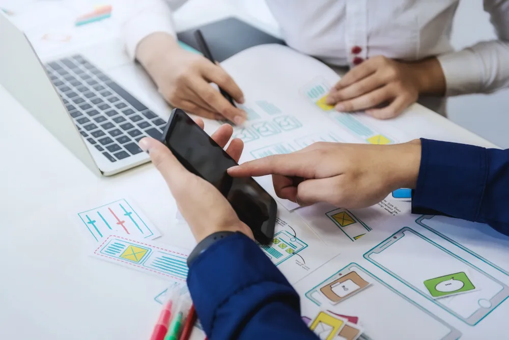 Three people examine a phone and interface mockups spread across a table