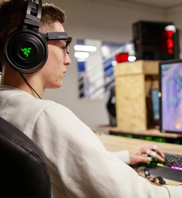 A young man wearing eye-tracking glasses, a headset and finger sensors, one hand on the keyboard, in front of a screen.