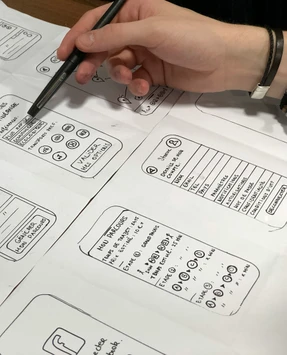 A hand points a marker at hand-drawn mobile app sketches on paper, including a journey planner screen.