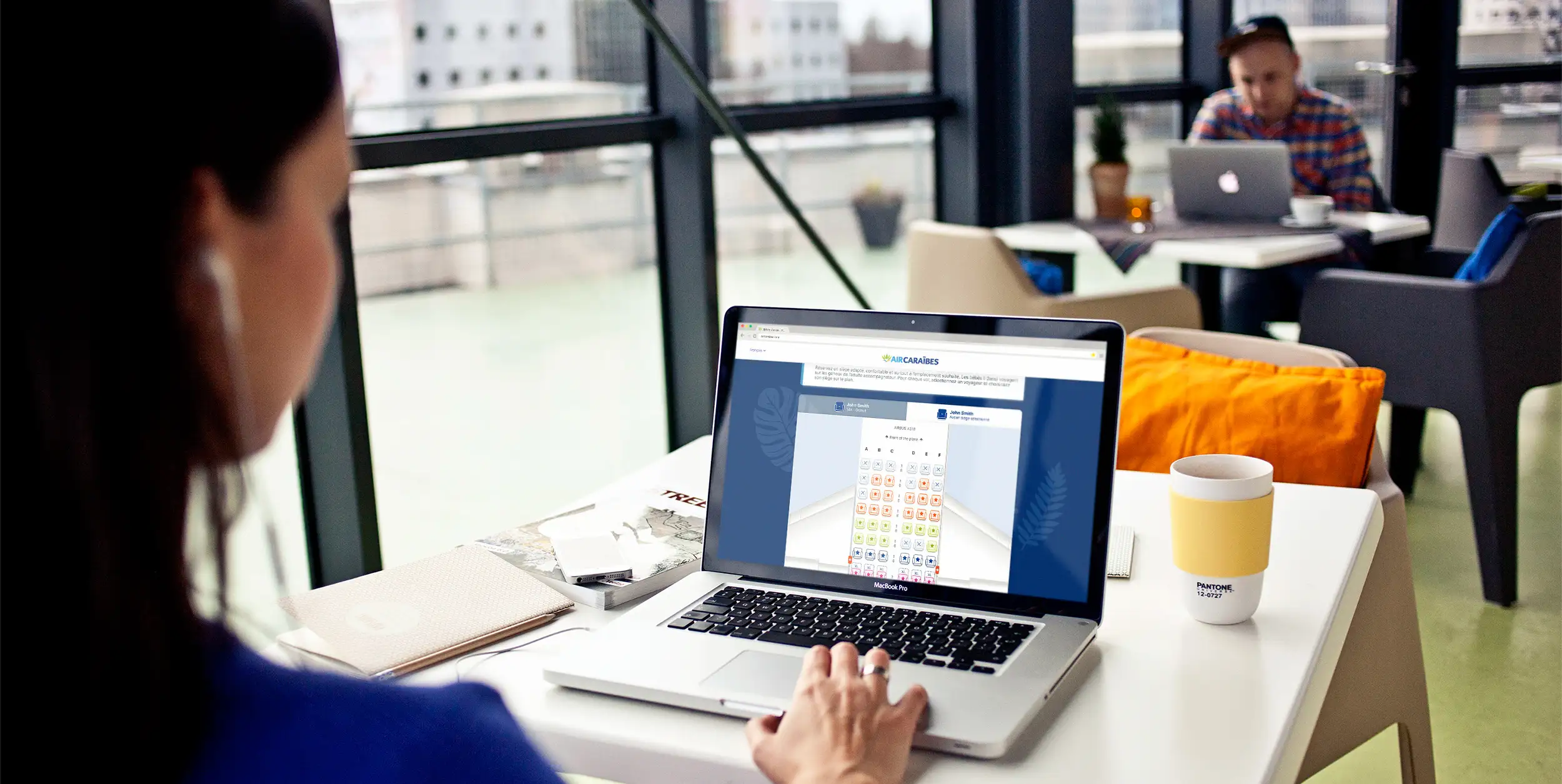 A person seen from behind in a glass-walled workspace, in front of a laptop open on a seat selection grid of the Air Caraïbes site.