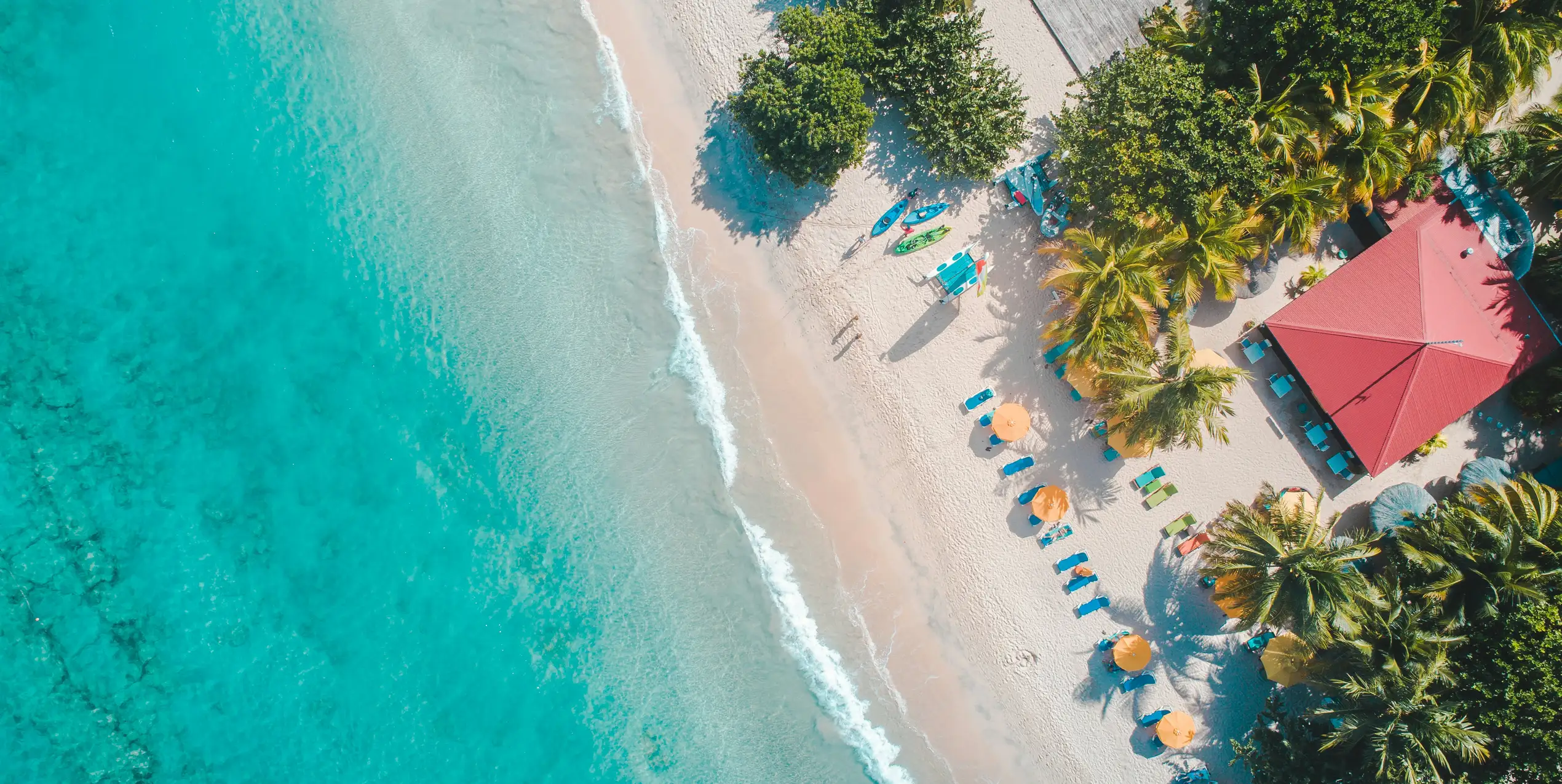 Aerial view of a white sand beach lined with coconut palms, with its parasols and a red-roofed building, beside turquoise water.