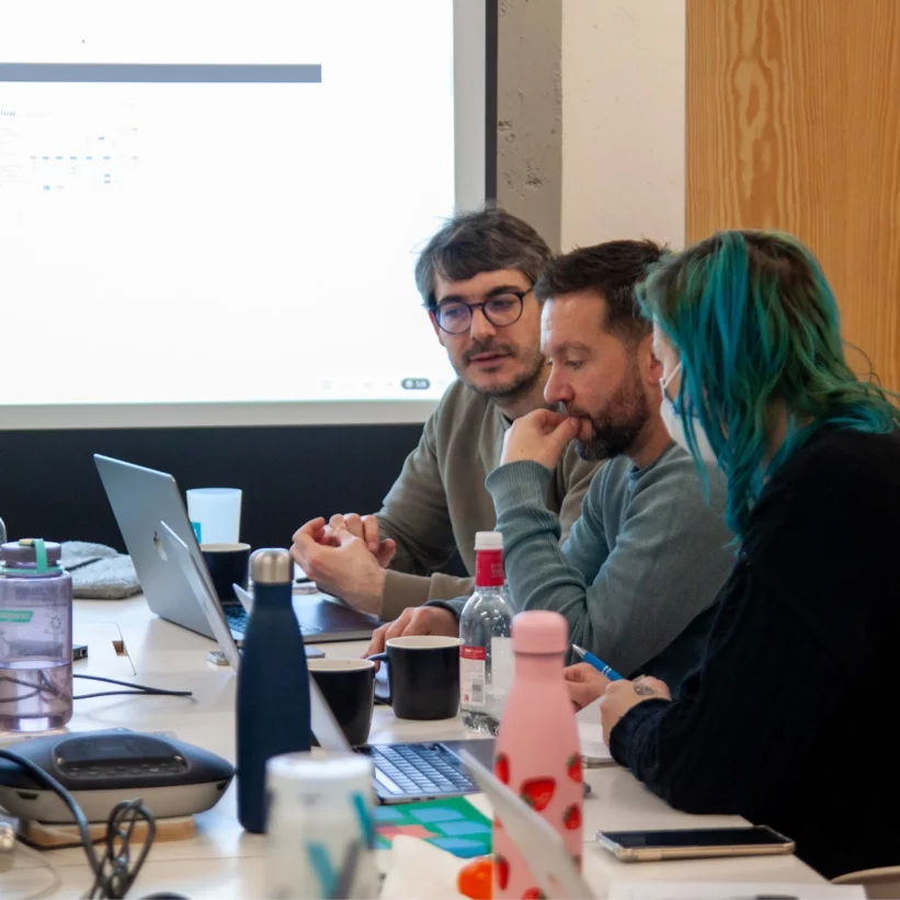 Trois personnes sont assises autour d'une table de réunion, devant un écran de projection affichant un diagramme de flux utilisateur (User Flow). Elles sont en pleine discussion, visiblement concentrées sur un appel d'offres. La table est encombrée de bouteilles d'eau, de gourdes, de tasses, d'un ordinateur portable et d'autres équipements de travail
