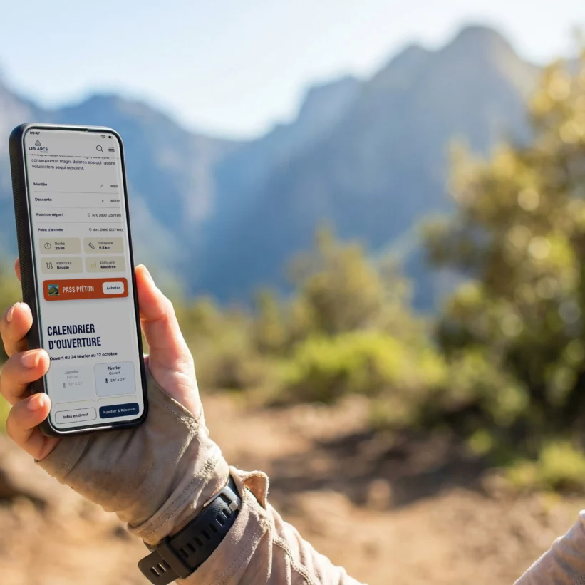 Une personne tient un smartphone affichant une application de randonnée avec un bouton Pass Piéton et un calendrier d'ouverture, sur fond de montagnes ensoleillées.